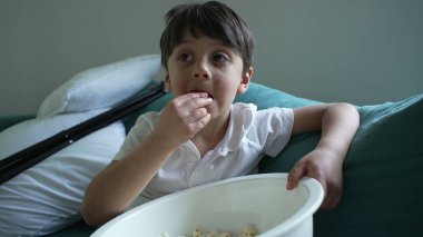 Küçük çocuk kanepede patlamış mısır yiyor, kendini bir şeyler izlemeye kaptırmış, çocukluğunun samimi bir anını yakalıyor.