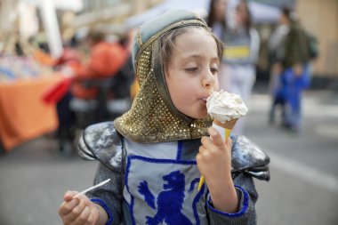 Ortaçağ şövalyesi kostümü giymiş bir çocuk açık hava festivalinde dondurma külahının tadını çıkarıyor. Eğlenceli sahne, tarihi canlandırma ile günlük eğlenceyi birleştirir. Çocuğun zevkini gözler önüne serer.