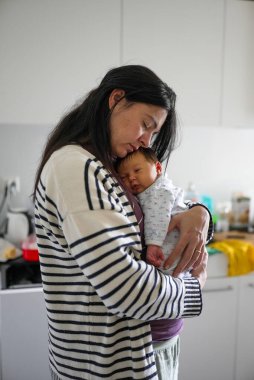 Anne uyumakta olan bebeğini nazikçe kucaklıyor. Huzurlu bir anda derin bir sevgi ve ilgi gösteriyor. İkisi de rahat ve rahat. Yakın bir aile bağını vurguluyorlar..