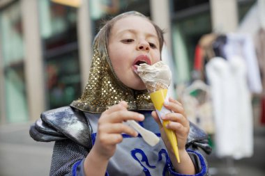 Ortaçağ şövalyesi kostümü giymiş bir çocuk açık hava festivalinde dondurma külahının tadını çıkarıyor. Eğlenceli sahne, tarihi canlandırma ile günlük eğlenceyi birleştirir. Çocuğun zevkini gözler önüne serer.