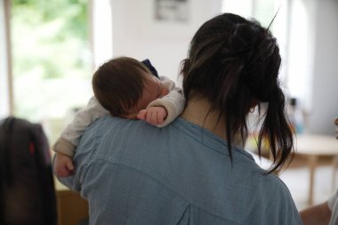 Anne, uyuyan bebeği omzunda tutuyor, yakın plan, mavi yakalı beyaz tulumlu bebek, kapalı mekan sahnesi, annelik bağı, rahatlatıcı an, ev ortamı, ebeveyn bakımı.