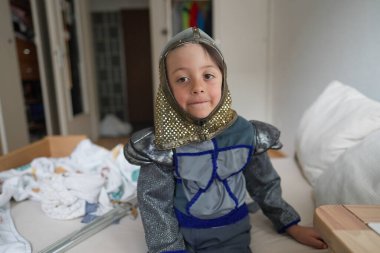 Küçük çocuk oyuncak kılıçlı bir şövalye gibi giyinmiş, yatakta oturuyor, yaratıcı bir oyun, ortaçağ kostümü giyiyor, yatak odası sahnesi, çocukluk, yaratıcılık, neşeli bir ifade, şövalye zırhı giyiyor.