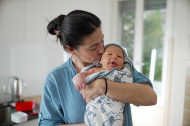 Anne yeni doğmuş bebeğini parlak bir mutfakta yumuşak bir battaniyeye sarar, sevgi dolu ve samimi bir anı yakalar, anneliğin sıcaklığını ve sevgisini yansıtır.