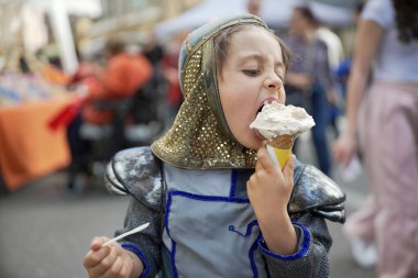 Ortaçağ şövalyesi kostümü giymiş bir çocuk açık hava festivalinde dondurma külahının tadını çıkarıyor. Eğlenceli sahne, tarihi canlandırma ile günlük eğlenceyi birleştirir. Çocuğun zevkini gözler önüne serer.