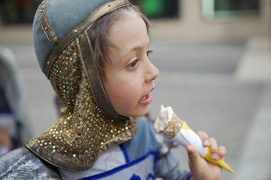 Ortaçağ şövalyesi kostümlü bir çocuk festivalde vanilyalı dondurma yiyor. Yan profil, çocuğun zevkini ve tarihi kıyafetlerin modern bir ziyafetle harmanlanmasını gösteriyor.