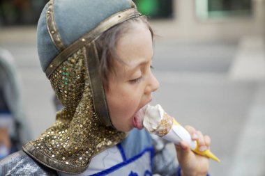 Ortaçağ şövalyesi kostümlü bir çocuk festivalde vanilyalı dondurma yiyor. Yan profil, çocuğun zevkini ve tarihi kıyafetlerin modern bir ziyafetle harmanlanmasını gösteriyor.