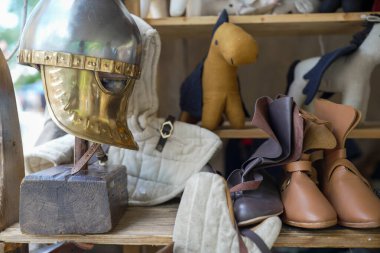 Ortaçağ miğferi ve diğer dönem aksesuarları, deri botlar ve kumaş eldivenlerle çevrili ahşap bir rafta, tarihi bir yeniden canlandırma etkinliğinde giysi ve zanaatkarlığı temsil ediyor.