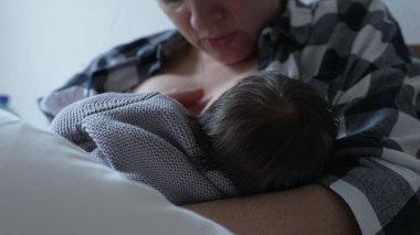 Anne, yeni doğan bebeği rahat gri bir battaniyeyle emziriyor, yakın bağ ve ilgiyi vurguluyor, anne ve çocuk arasındaki besleyici ilişkiyi gösteriyor ve emzirmenin önemini anlatıyor.