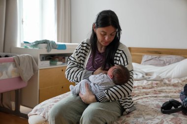 Anne sevgiyle yeni doğan bebeğini bir yatak odasında tutuyor, annelik bakımını ve bağ kurmayı ifade ediyor. Arka planda bebekle ilgili eşyalar olan rahat, rahat bir ortam..