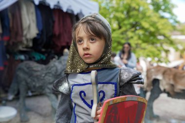 Çocuk şövalye gibi giyinmiş, ciddi görünüyor, bir ortaçağ festivalinde kılıç ve kalkan tutuyor. Sahne, tarihi canlandırma atmosferini ve çocuğun odaklanmış ifadesini yakalıyor.