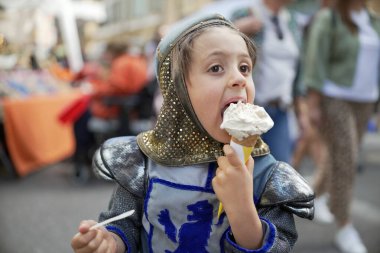 Ortaçağ şövalyesi kostümü giymiş bir çocuk açık hava festivalinde dondurma külahının tadını çıkarıyor. Eğlenceli sahne, tarihi canlandırma ile günlük eğlenceyi birleştirir. Çocuğun zevkini gözler önüne serer.