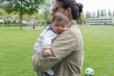 Anne parkta bebeği kollarında tutuyor, beyaz tulumlu bebek, güneş gözlüklü ve yeşil ceketli anne, arka planda futbol golü, açık hava sahnesi, anne bakımı, aile gezisi.