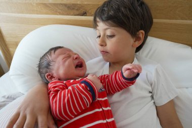 Büyük kardeş beşikte kırmızı çizgili tulumlar içinde, her ikisi de yatakta dinleniyor, aile ortamında kardeş bakımını ve bağlanmayı tasvir ediyor, sevgi ve bağlantıyı vurguluyor.