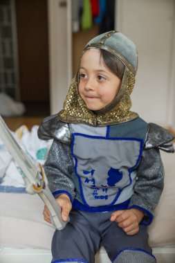 Şövalye kostümlü genç adam yatakta oturuyor, yaratıcı oyun, ortaçağ temalı kıyafet, çocukluk eğlencesi, mutlu ve oyuncu, ev ortamı, yaratıcı oyun, gülümseyen çocuk, oyuncak kılıç