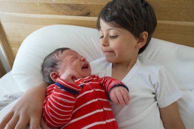 Büyük kardeş kırmızı bebek tulumuyla yatakta ağlıyor koruyucu ve şefkatli bir ilişki gösteriyor sıcak bir aile ortamında kardeş sevgisinin ve şefkatinin altını çiziyor.