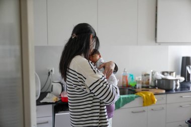 Anne, uyuyan bebeğini mutfakta beşikte besliyor ve ilgi gösteriyor. Arka planda ev eşyaları ve mutfak gereçleri var..