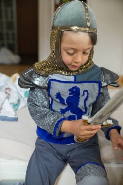Şövalye kostümlü genç adam yatakta oturuyor, yaratıcı oyun, ortaçağ temalı kıyafet, çocukluk eğlencesi, mutlu ve oyuncu, ev ortamı, yaratıcı oyun, gülümseyen çocuk, oyuncak kılıç