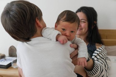 Küçük çocuk, annesinin kucağında otururken yeni doğan kardeşini kucaklıyor. Yürek ısıtan bir aile sevgisi ve ebeveyn bakımı anı sergiliyor. Aile ve çocukluk teması için mükemmel.