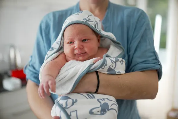 stock image Mother holding newborn baby wrapped in a blue and white animal print hooded towel, both smiling, baby looking curious and content, mother wearing a light blue shirt, bright kitchen background