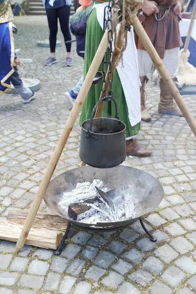 Ortaçağ yemek düzeneği, ateş çukurunda asılı duran bir tencere, kaldırım taşlarıyla çevrili, arka planda dönemin kostümlü insanları, otantik bir tarihi atmosfer yaratıyor.