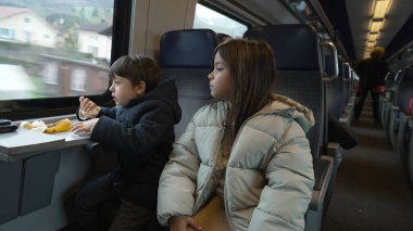 Trende gezen, muz yiyen ve pencereden dışarı bakan, manzaranın tadını çıkaran çocuklar, sıcak ceketli kardeşler, yolculukta atıştırmalık bir şeyler yiyorlar.