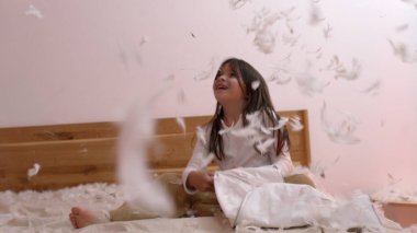 Mutlu kız yatak odasında tüyleriyle yastık savaşından zevk alıyor etrafta uçuyor eğlenceli bir ortam yaratıyor beyaz tişörtlü ve bej pantolonlu çocuk gülüyor