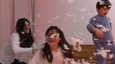 Neşeli çocuklar ve anne şakacı yastık savaşı yapıyor, pembe perdelerle bir odada uçuşan tüyler, eğlenceli ve enerjik bir atmosfer yaratıyor, aile bağlarını ve neşeli anları ön plana çıkarıyor.