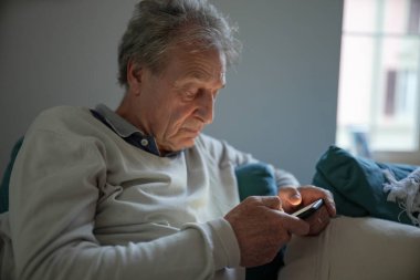 Yaşlı bir adam akıllı telefonuna odaklanmış, ev ortamında bir kanepede oturuyor. Sahne, teknolojinin günlük yaşamla bütünleşmesini ve kullanıcının konsantrasyonunu vurgular..