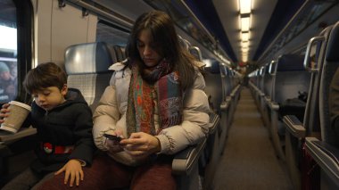 Anne ve oğlunun birlikte oturduğu bir trendeki aile anı, akıllı telefonuna ve elinde bir kupa tutan oğluna odaklandı, seyahat sırasında bir teknoloji ve bağlanma karışımı yakaladı.