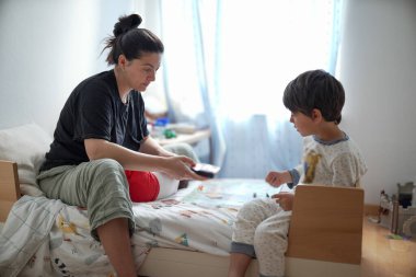 Anne ve oğul, çocuğun yatağında, elinde bir şeye odaklanmış bir şekilde etkileşim halindeler. Sahne, aile ortamındaki samimi bir anı tasvir ediyor, bağlarını vurguluyor..