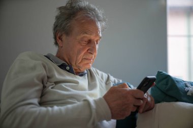 Kanepede otururken akıllı telefon kullanan yaşlı bir adam faaliyetlerine dalmış. Sahne, yaşlılar arasında modern teknolojinin rahat bir ev ortamındaki kullanımını gösteriyor..