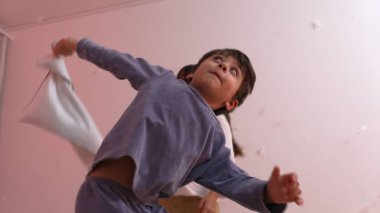 Pijamalı enerjik çocuk, bir kızla oynaştığı yastık kavgasında eğleniyor, dinamik hareketler ve heyecanı ev ortamında gösteriyor.