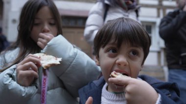 Açık havada pizzayı seven iki çocuk, halka açık bir ortamda insanlarla çevrili, paylaşılan bir anın sıcaklığını ve zevkini vurguluyorlar.