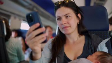 Annem akıllı telefonu trende uyuyan yeni doğmuş bebeği beşikte tutarken tutuyor, telefonu odaklanmış bir ifadeyle kontrol ediyor. Bebek kucağında huzur içinde uyuyor.