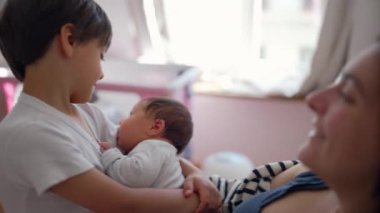 Anne, büyük oğlunun bebek kardeşini kucağına alışını, neşeli aile etkileşimini sıcak bakımı, sıcak ev ortamını, kardeş bağını, sevgi dolu bağı, doğal ışık, erken çocukluk.