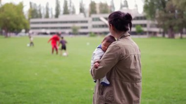 Anne kucağında uyuyan bir bebek, arka planda insanlarla parkta duruyor, kamu düzeninde bakım ve bakımı ön plana çıkarıyor.