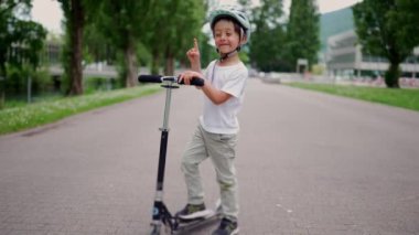 Genç çocuk sessiz park yolunda, gür ağaçlar ve dingin çevre ile çevrili, açık hava keşfi ve çocukluk aktivitelerini vurgulayan scooter 'da duruyor.