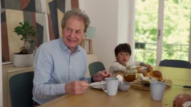 Büyükbaba yemek masasında sıcacık gülümsüyor, küçük torunuyla kahvaltısını yapıyor, neşeli ve hassas bir aile anı, sevgi, bağ ve birliktelik teması.