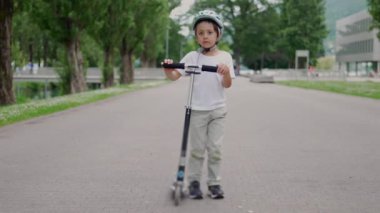 Kask takan küçük bir çocuk parktaki kaldırımlı yolda scooterla duruyor, çevresi yeşilliklerle çevrili, açık hava aktivitelerine ve güvenliğe odaklanıyor.