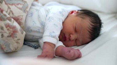 Yeni doğmuş bebek, yumuşak tulum giymiş bir yatakta huzur içinde uyuyor. Yavrunun sakin ifadesi ve rahat duruşu rahat bir ortamda huzurlu bir uyku anı yakalar.