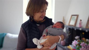 Büyükannem yeni doğmuş bir bebeği gururlu bir gülümsemeyle kucağında tutuyor, sıcak bir yerde duruyor, aile sevgisini ve çoklu-kuşak bağını gösteriyor.