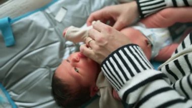 Bebek bakıcı tarafından yumuşak bir paspasın üzerinde nazikçe giydiriliyor. Bebek sakince yukarı bakar. Bakıcının elleri elbiseyi özenle ve şefkatle ayarlar.