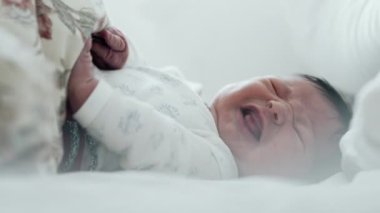Yeni doğmuş bebek yatakta ağlıyor, rahatsız edici bir şekilde elini kaldırıyor. Bebeğin yüzünde sıkıntı var, ve jestler tedirginliği gösteriyor.