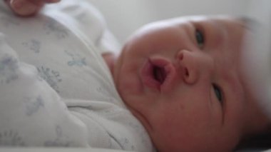 Yeni doğmuş bebek yatakta yatıyor, gözleri sonuna kadar açık, merakla etrafı keşfediyor. Bebek yüzü merak ve ilgi gösteriyor, doğal, yumuşak ışık içeride.