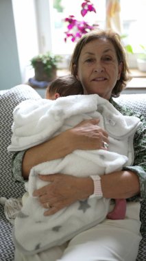 Grandmother tenderly kissing her swaddled grandbaby, both wrapped in white blankets, seated on a patterned couch in a well-lit home environment clipart