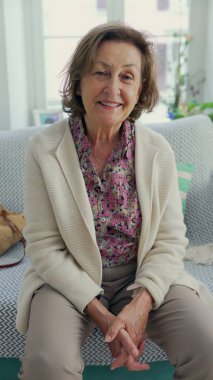 Yaşlı bir kadın, parlak ve rahat bir oturma odasında, kanepede otururken sıcak bir şekilde gülümsüyor, çiçekli bir gömlek ve açık bir hırka giyiyor, hoş bir atmosfer yaratıyor.