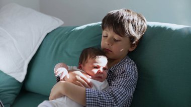 Küçük çocuk, yeni doğan kardeşini sevgiyle kanepede tutuyor ve sıcak bir yuva kuran kardeşler arasındaki hassas ve koruyucu bağı vurguluyor.