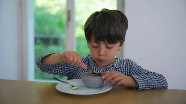 Mavi kareli gömlekli genç adam kahvaltılık gevrek yiyor, yeşil yapraklı arka planı pencereden görülebilen modern bir mutfakta yemeğine odaklanıyor.