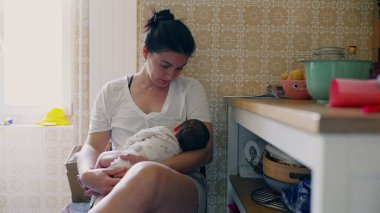 Anne yeni doğmuş bebeğini rahat bir mutfak ortamında nazikçe kucaklıyor ve besliyor, erken yaşta annelik, ebeveynlik bağı ve bebek bakımını sıcak, rahat bir ortamda gösteriyor.