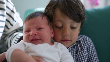 Küçük bir çocuk ağlayan yeni doğan bir kardeşi rahatlatıyor, sıcak bir yuvadaki kardeşler arasındaki koruyucu ve sevgi dolu bağı gösteriyor.